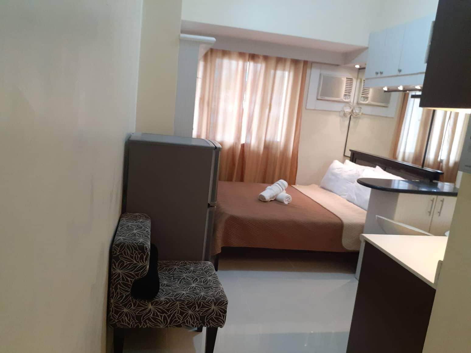 A compact hotel room with a bed, a small desk, a chair, and a kitchenette.
