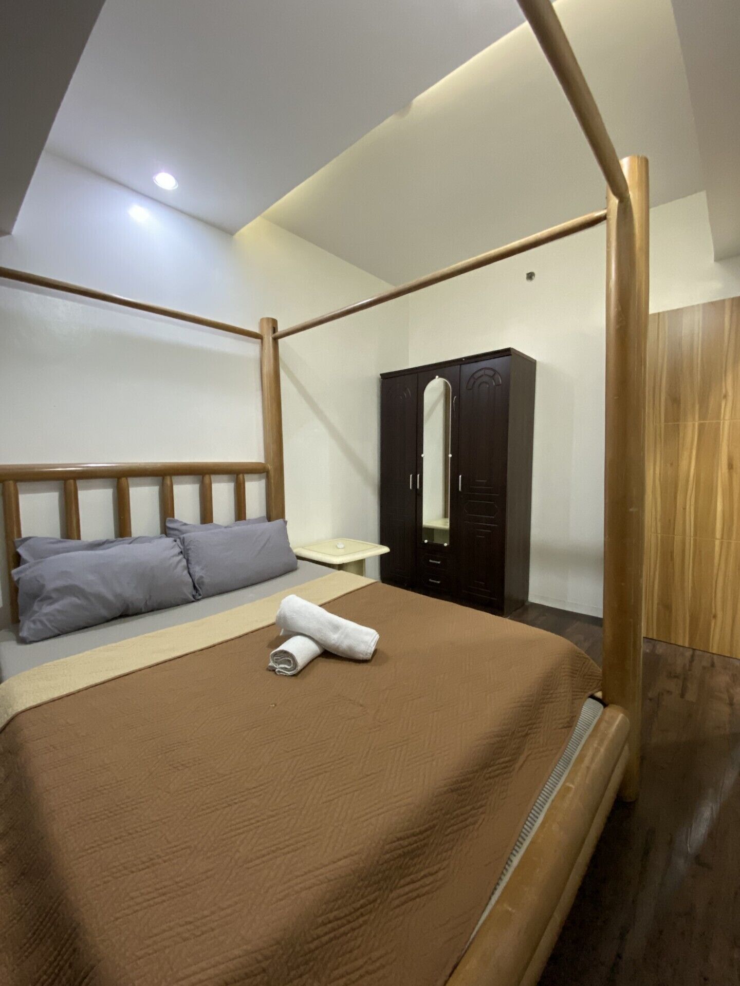 A modern hotel room with a four-poster bed, a wooden wardrobe, and a small bedside table.