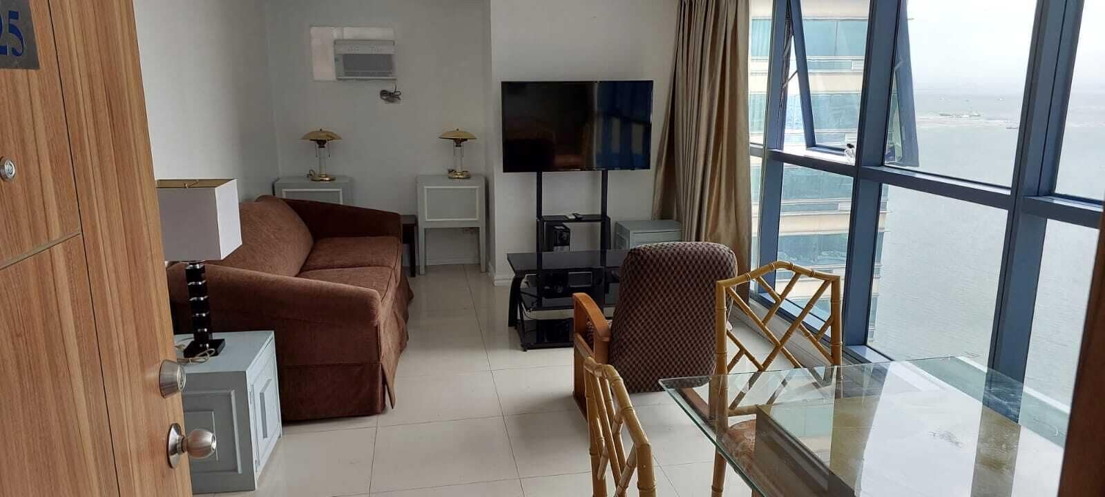 Ένα σύγχρονο δωμάτιο ξενοδοχείου με ένα μεγάλο παράθυρο που προσφέρει θέα στη θάλασσα, έναν καναπέ, ένα μικρό τραπέζι και μια τηλεόραση.