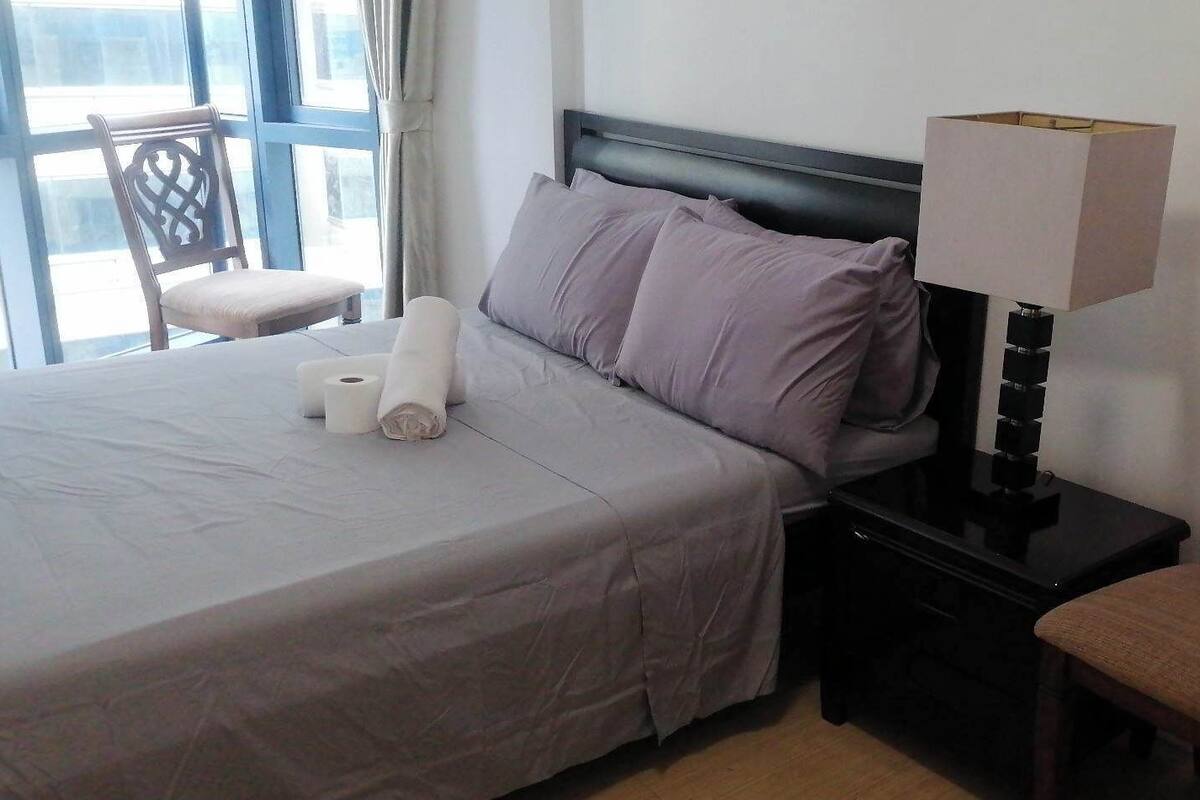 A neatly made bed with a grey bedspread, two pillows, and a rolled-up towel. There is a bedside table with a lamp and a chair with a wooden backrest.