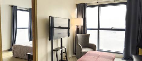 Een hotelkamer met een bed, een televisie op een voet, een stoel, een tafel en een groot raam met gordijnen.