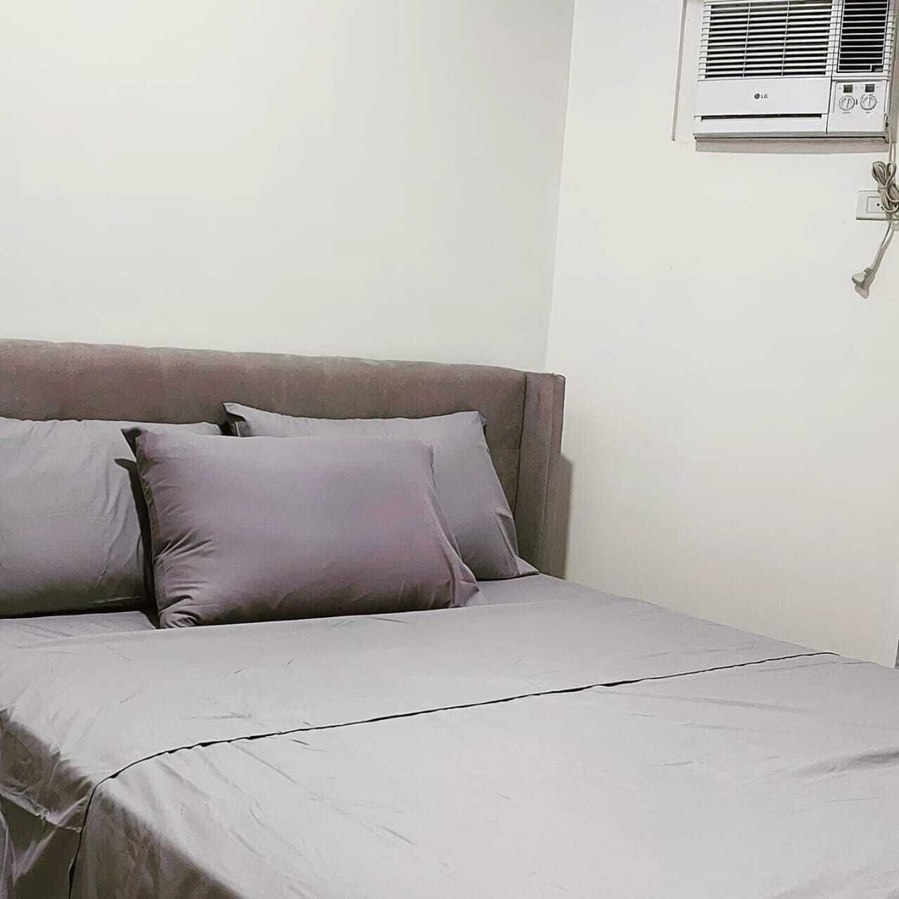 A bed with a grey mattress and pillows, a white wall, and an air conditioning unit mounted on the wall.