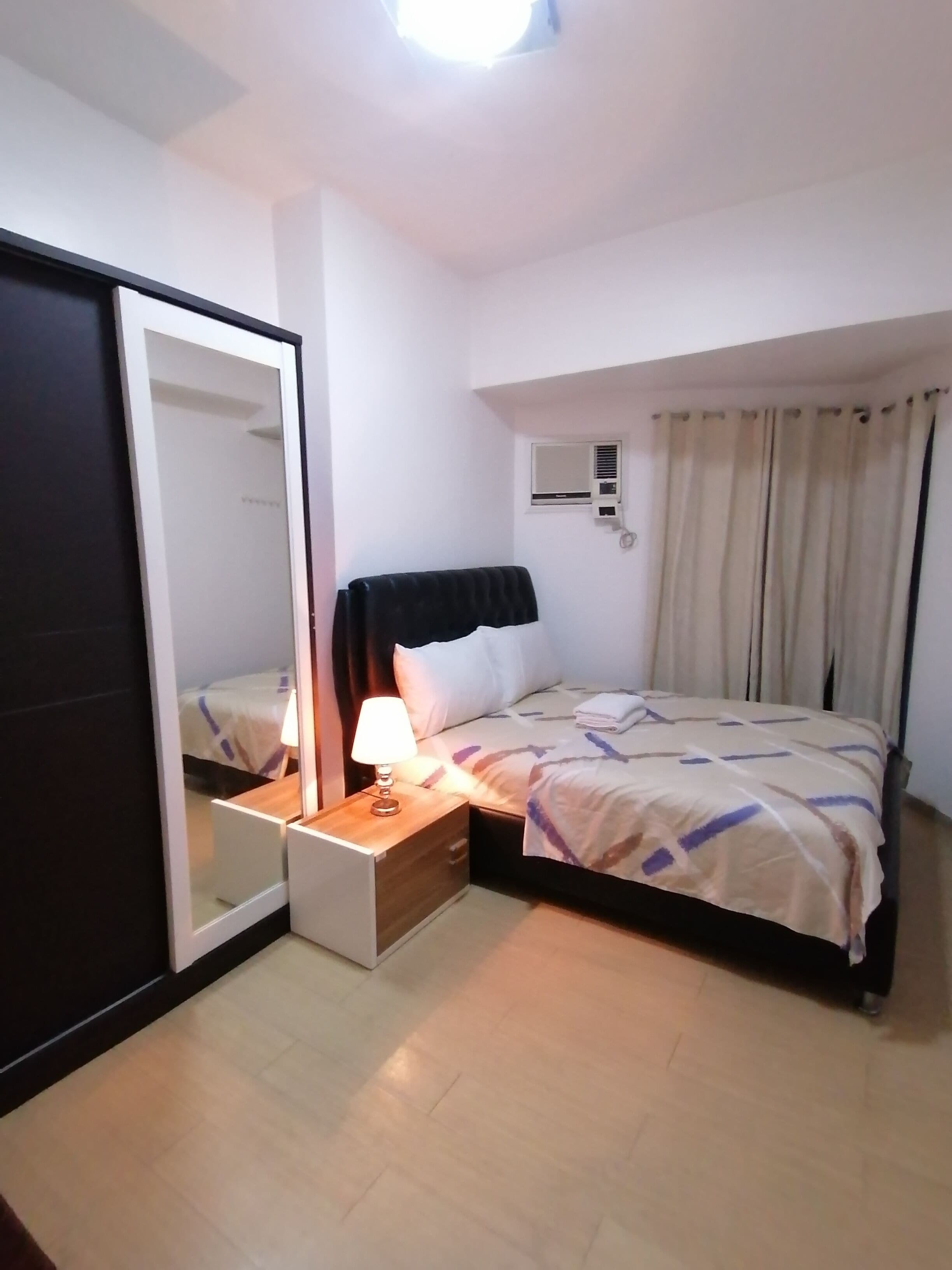 A hotel room with a bed, a bedside table with a lamp, a wardrobe, a window with curtains, and an air conditioning unit.
