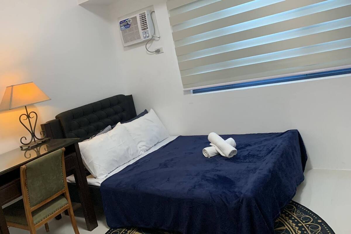 A hotel room with a bed, a desk with a lamp, a chair, and a window with blinds.