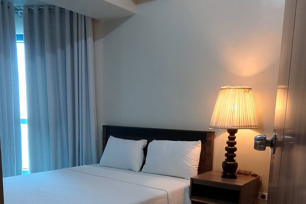 A neatly made bed with white linens, a wooden nightstand with a lamp, and a wooden floor.