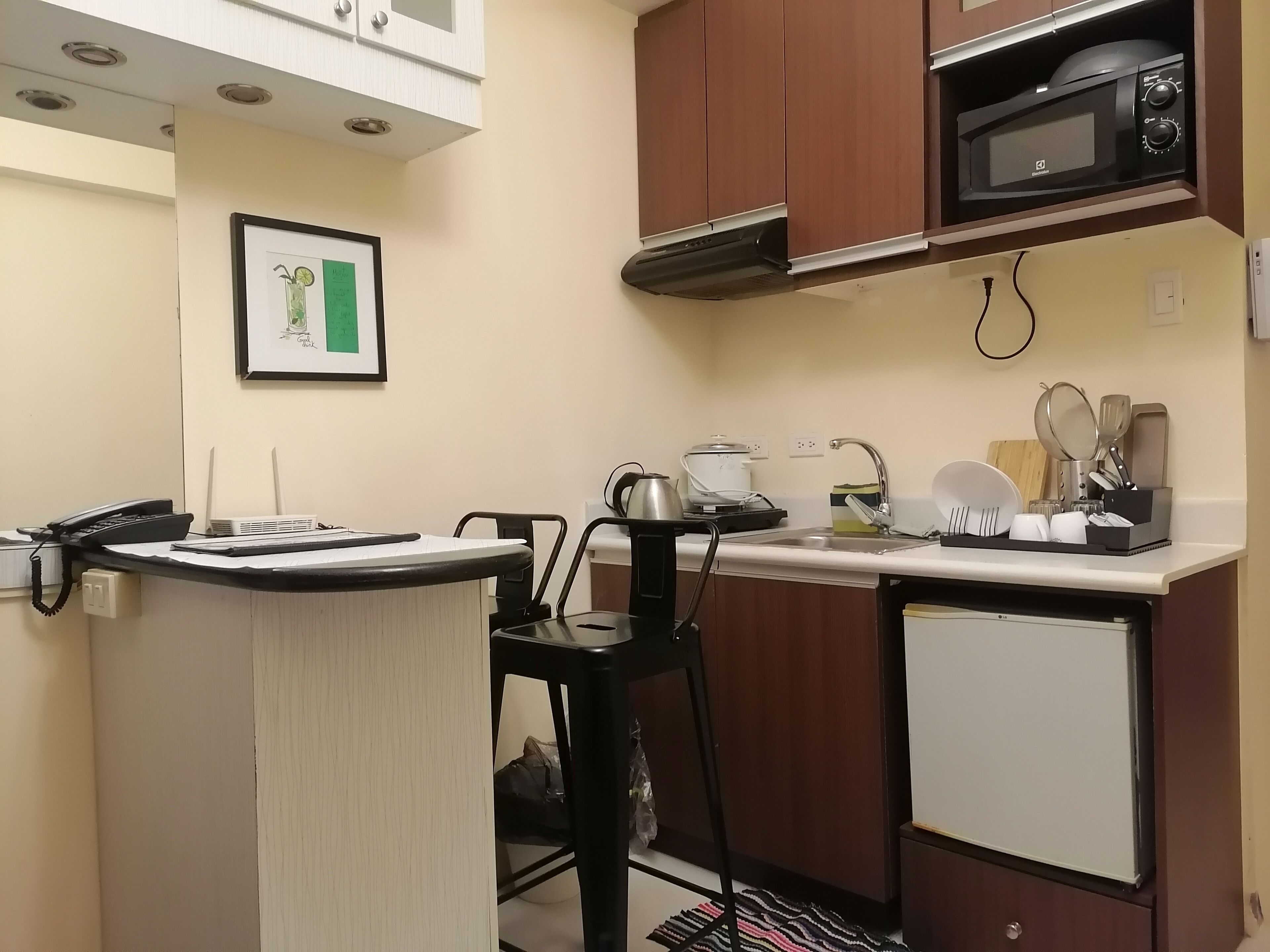 A compact kitchen with a microwave, kettle, and small refrigerator. There is a framed picture on the wall and a telephone on the counter.