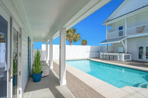 A covered outdoor swimming pool area with a white fence, a poolside bar, and a two-story building in the background.