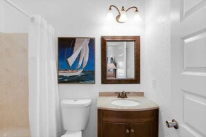 A bathroom with a white toilet, a sink with a wooden vanity, a painting of a sailboat, and a mirror with a wooden frame. - Epic! Ocean views! Sleeps 20! Pool! Golf cart! (Destin)