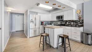 A modern kitchen with white cabinets, a central island with stools, stainless steel appliances, and a black tiled backsplash. - Gulf Shores Condo 403 Gorgeous 4th Floor Beachfront View (Port Aransas)