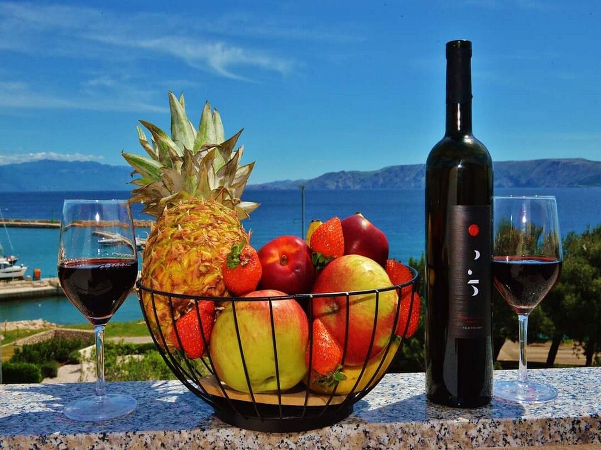 A fruit bowl with a bottle of wine and glasses on a table with a scenic view.