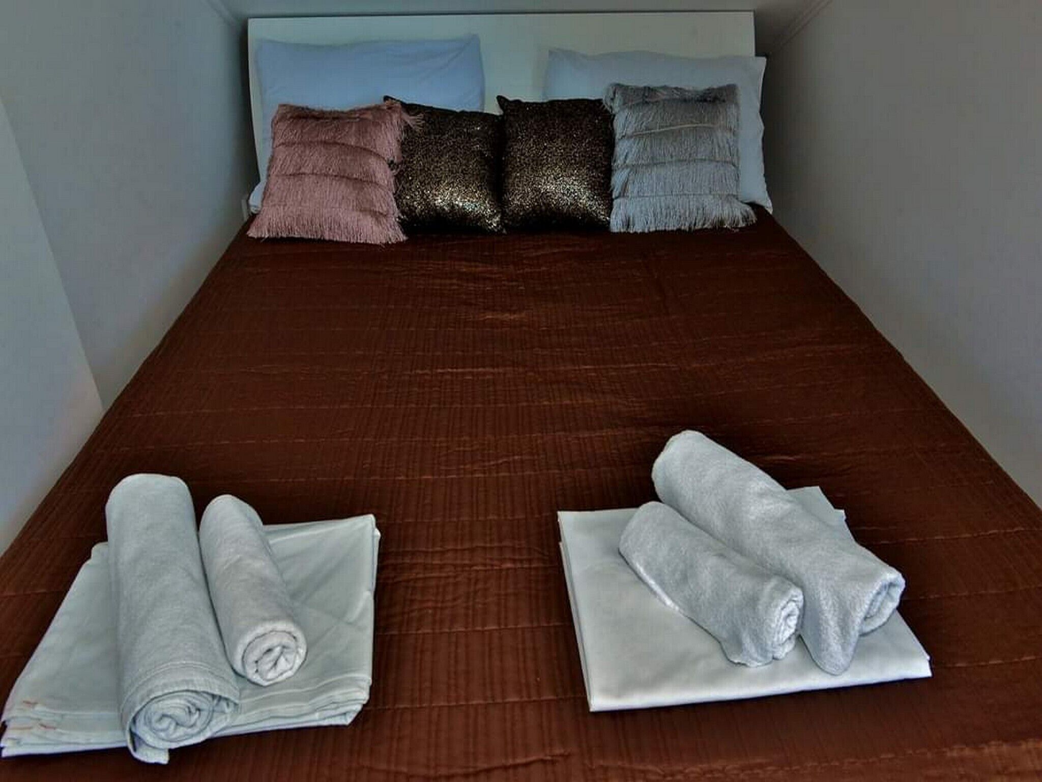 Cama bem arrumada com colcha marrom, dois toalhas enroladas e quatro travesseiros em cores e texturas variadas.