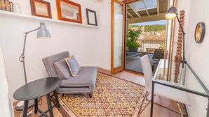 Ein gemütlicher Wohnbereich mit grauem Sofa, einem kleinen runden Tisch und einem Glastisch mit Stuhl. An der Wand befinden sich gerahmte Bilder und ein großes Fenster mit Blick auf Gebäude und Grünflächen.