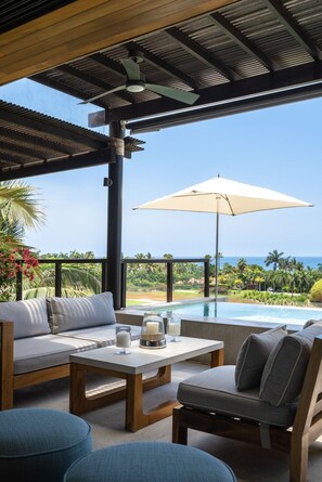 A poolside area with a covered seating area, a table with drinks, and a view of the ocean.