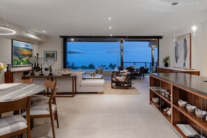 A modern living room with a dining area, large windows, and a view of the ocean.