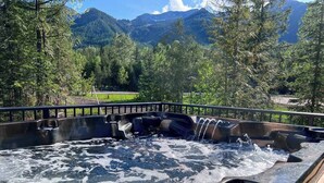 A hot tub with a wooden deck and a metal railing, surrounded by a forest and mountains.