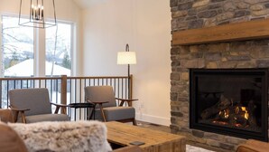 A cozy living room with a stone fireplace, two armchairs, a wooden coffee table, and a view of a snowy landscape through large windows.