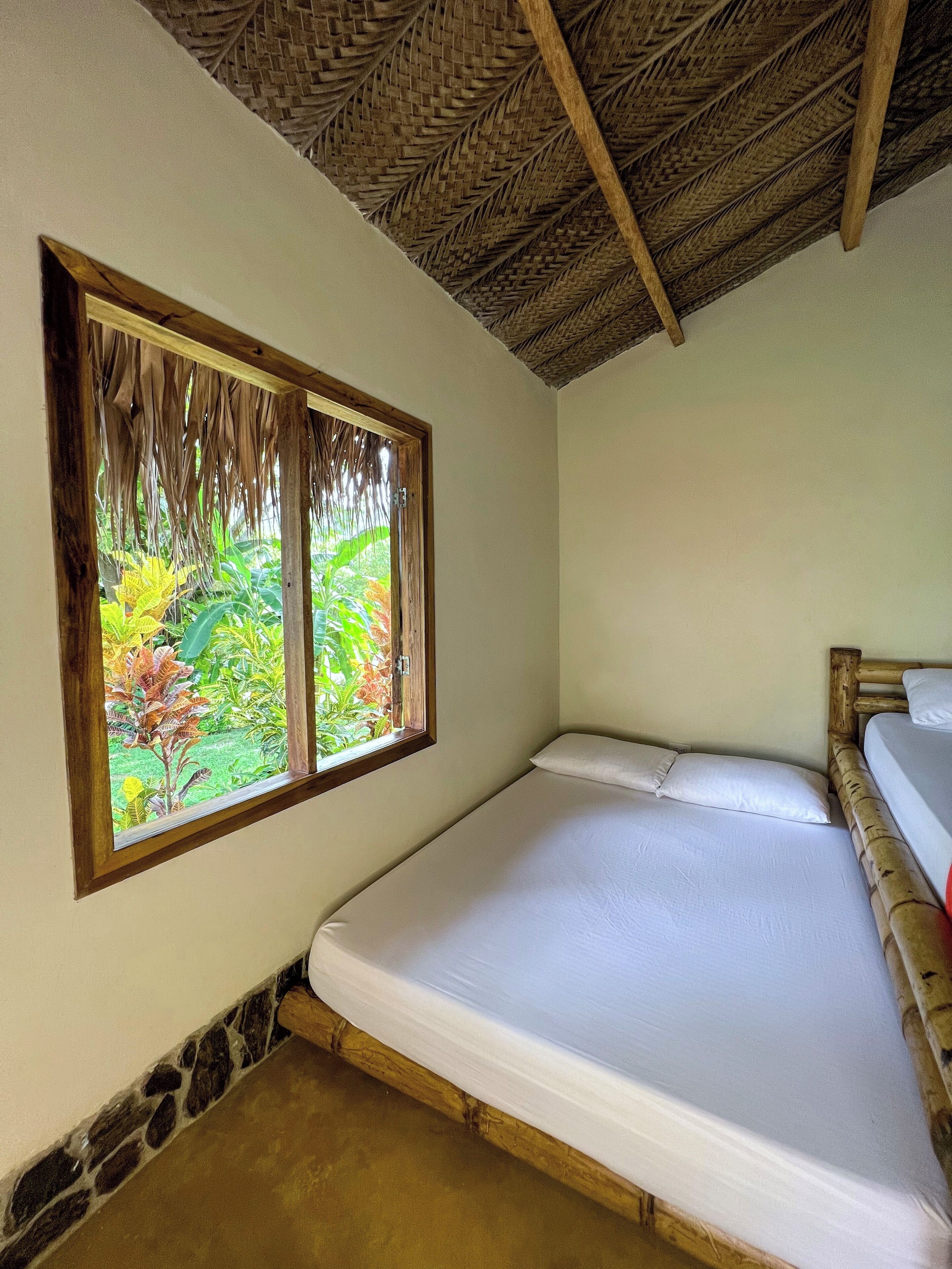 A single bed with a wooden headboard, a window with a view of greenery, and a thatched roof ceiling.