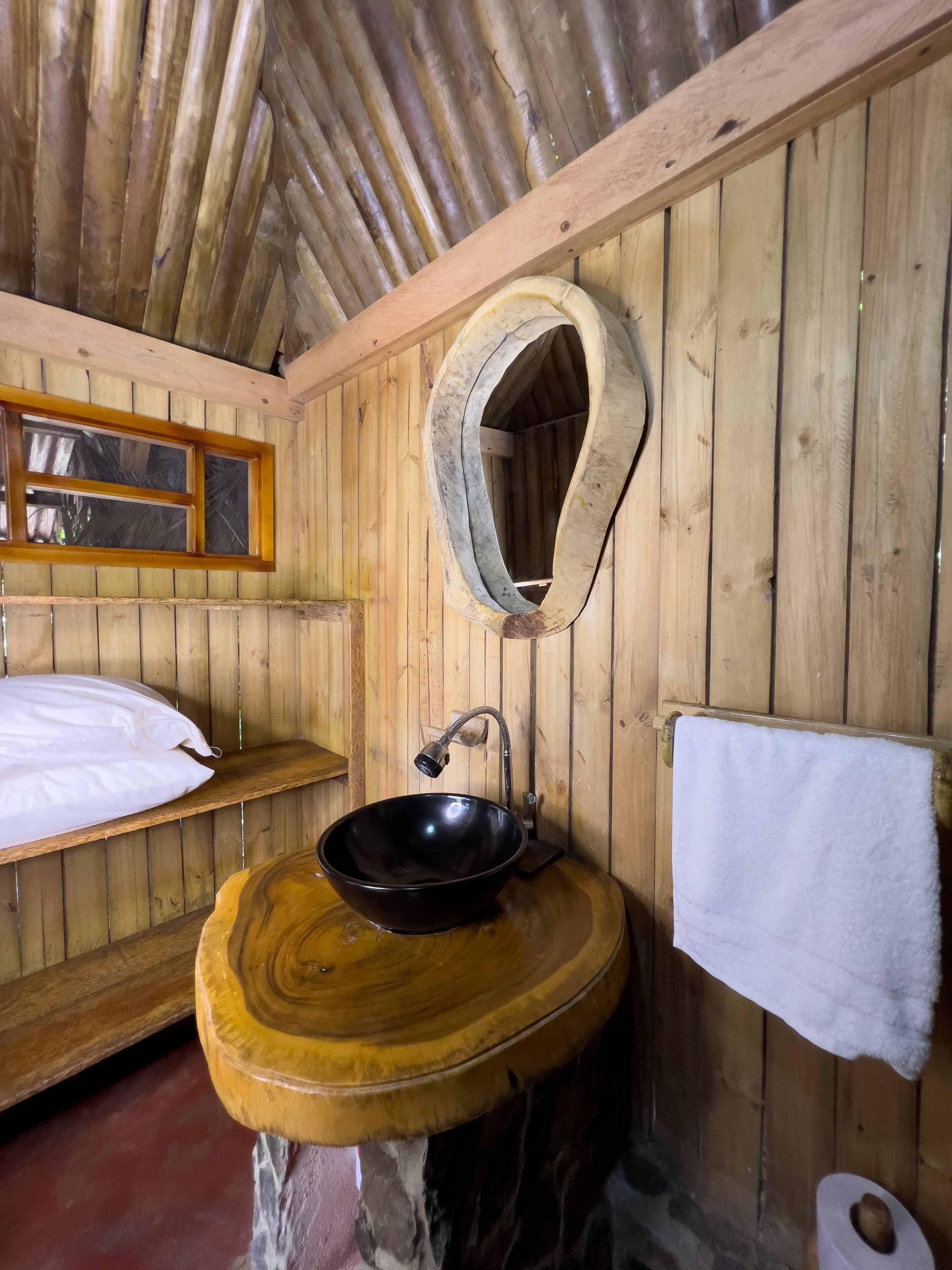 Interieur van een houten hutje met een ronde houten wastafel, een spiegel, een bed met witte lakens en een handdoek aan de muur.