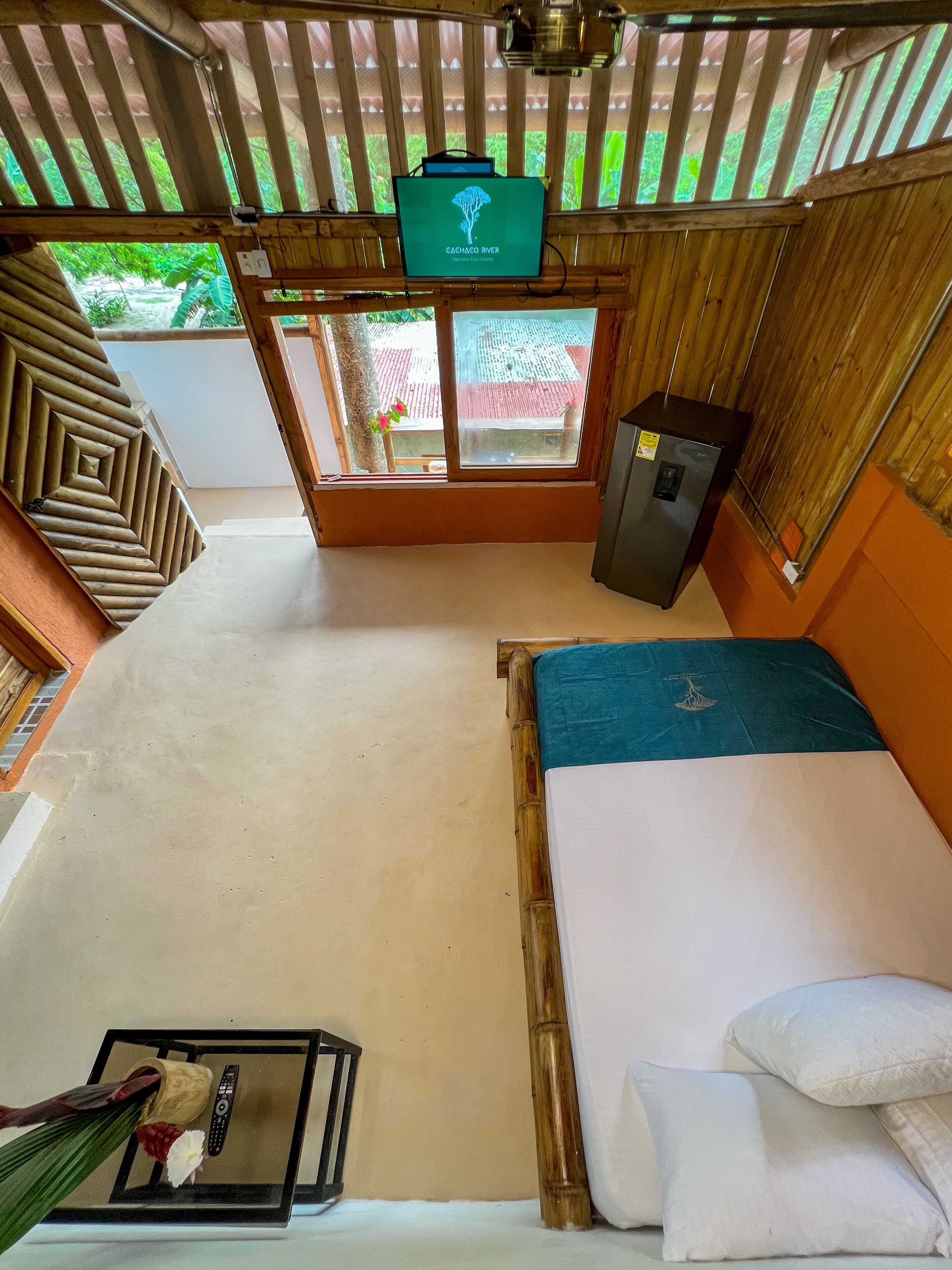 A room with a bed, a small table with a remote control, a television on the wall, and a window with a view of greenery and a building outside.