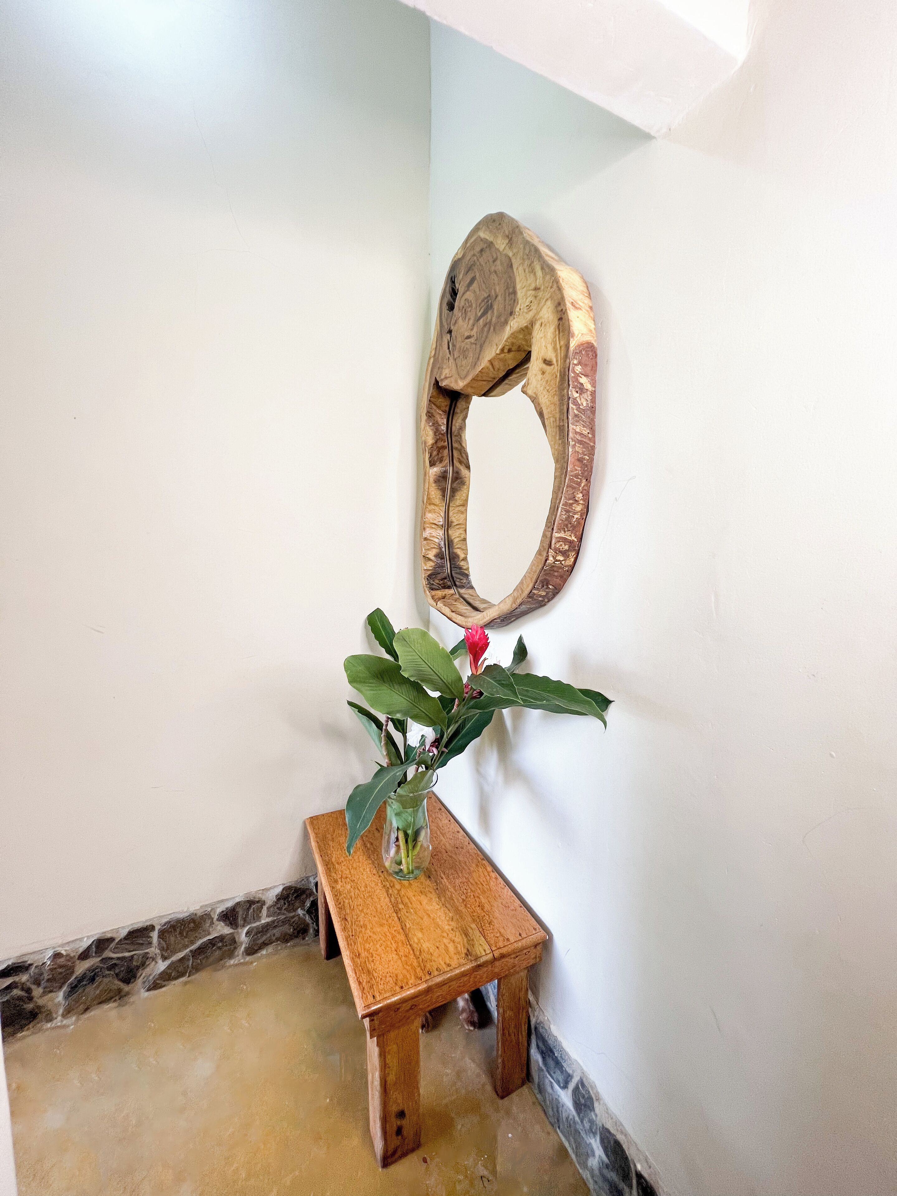 Una mesa de madera con un jarrón de plantas verdes y una flor roja, ubicada frente a una pared blanca con una escultura de madera.
