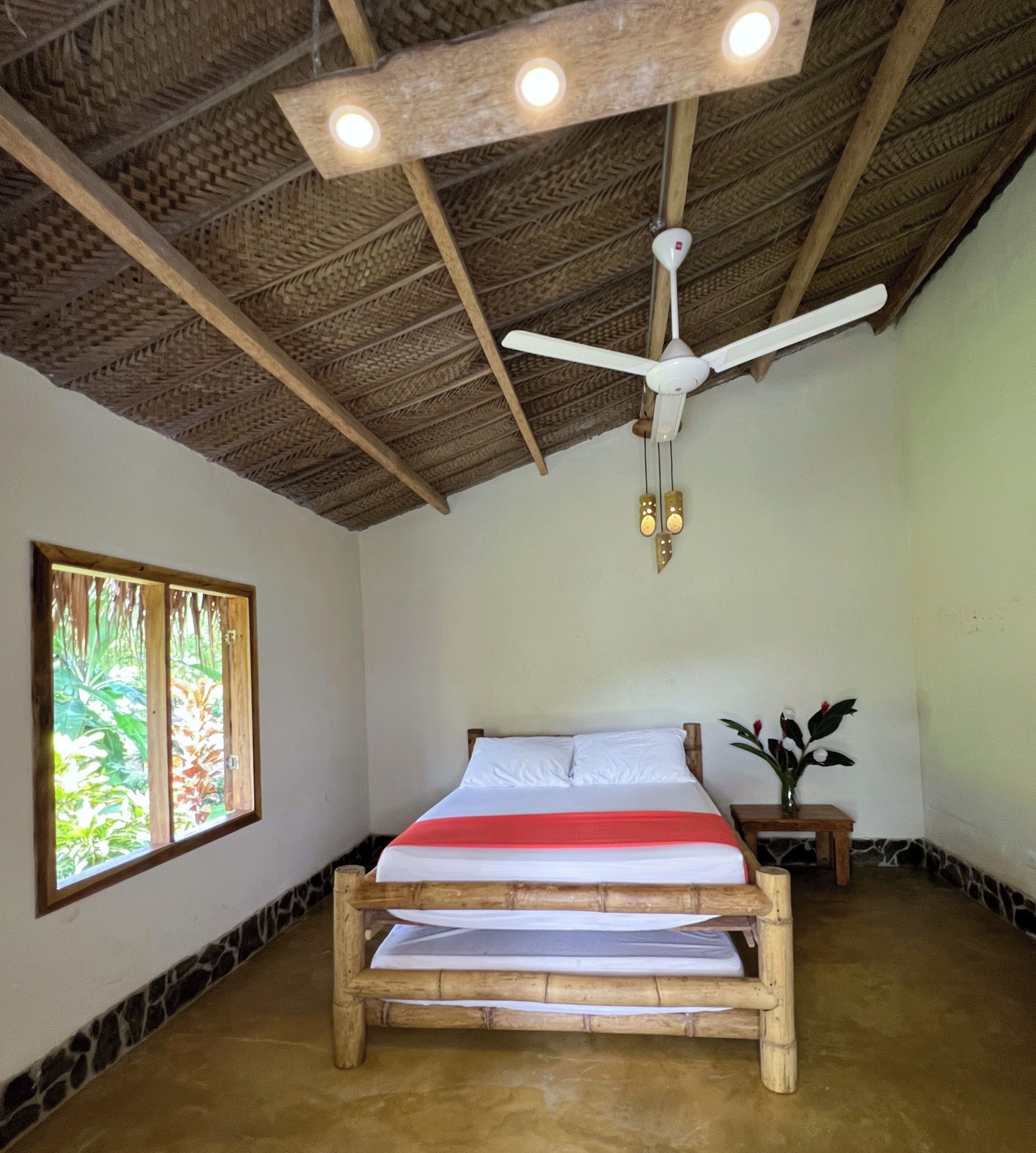 A room with a bed, a ceiling fan, a window with a view of greenery, and a small table with a vase.