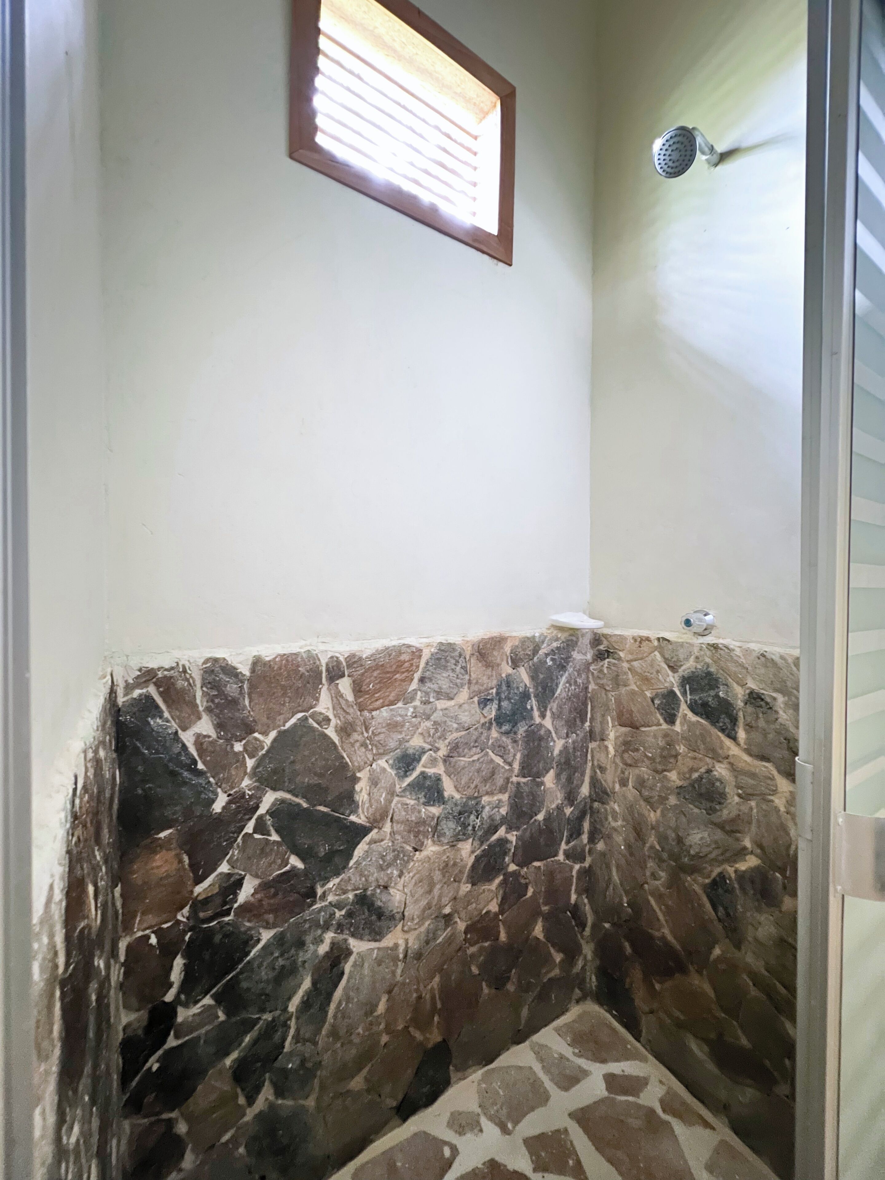 A bathroom with a stone wall, a showerhead, and a window with blinds.