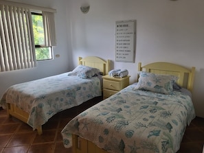 A room with two single beds, each with a matching headboard and footboard, a bedside table with a lamp, and a wall-mounted sign listing positive attributes of the house. - Beachfront Community Ocean View 2Br/2Ba in Los Almendros, Playa Ocotal (Playa Ocotal)