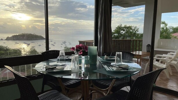 A dining area with a glass table and chairs, a vase with flowers, and a view of the sea and boats. - Beachfront Community Ocean View 2Br/2Ba in Los Almendros, Playa Ocotal (Playa Ocotal)