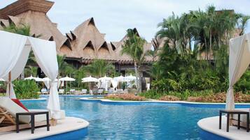A resort pool area with lounge chairs and umbrellas, surrounded by tropical vegetation and thatched-roof buildings.