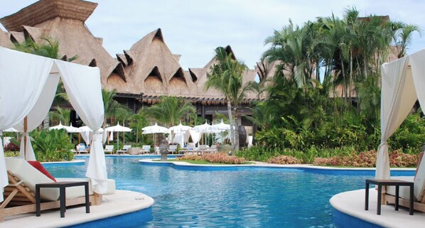 A resort pool area with lounge chairs and umbrellas, surrounded by tropical vegetation and thatched-roof buildings. - Vidanta Resort Grand Luxxe Deluxxe 1 bedroom in Playa del Carmen Mexico (Playa del Carmen)
