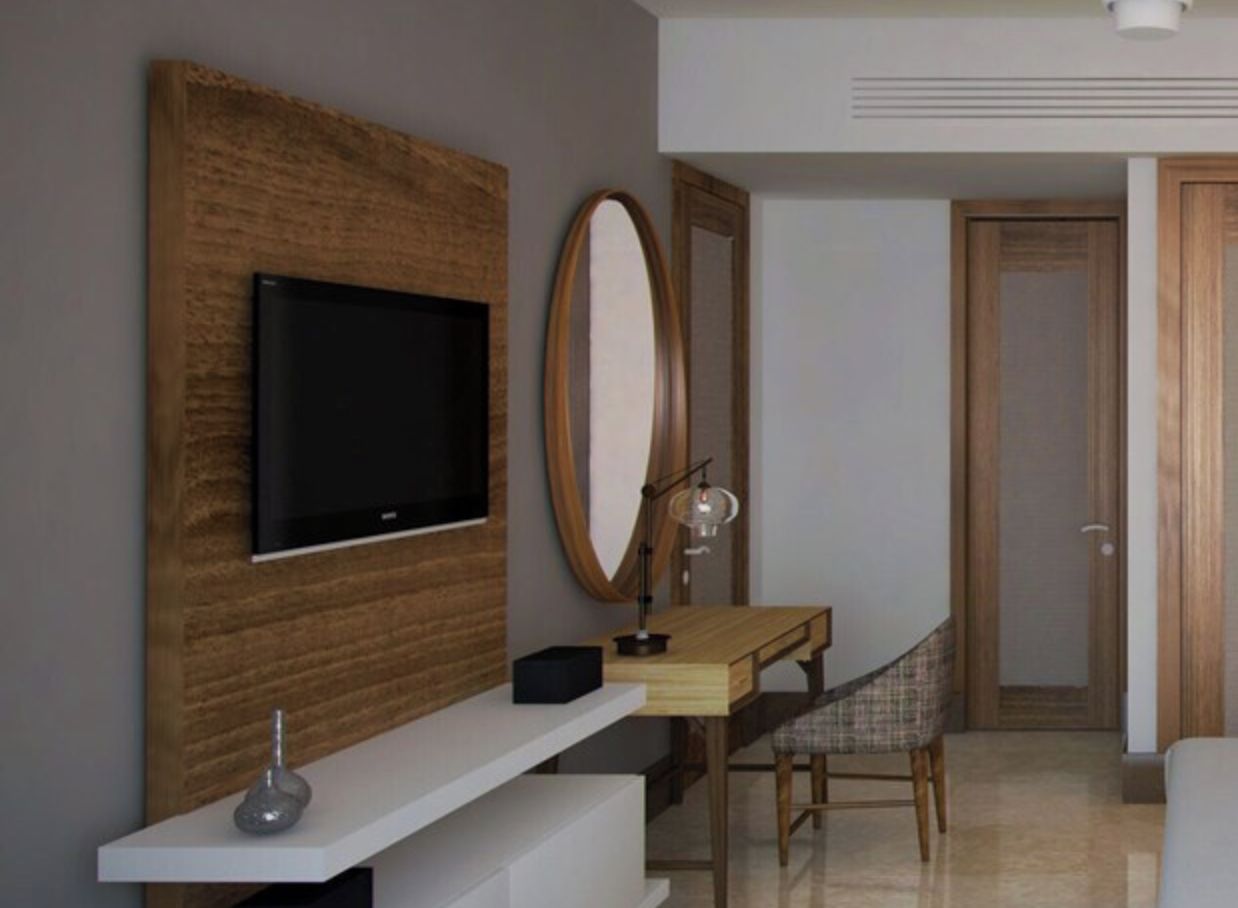 A modern hotel room with a flat-screen TV mounted on a wooden panel, a desk with a chair, a large mirror, and a door leading to another room.