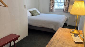 A small, single-bed room with a wooden nightstand, a bench, a window with floral curtains, and a wall-mounted heater.