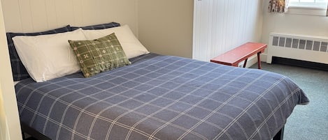 Un dormitorio con una cama, almohadas, un cuadro enmarcado, una mesita y una ventana con cortinas.
