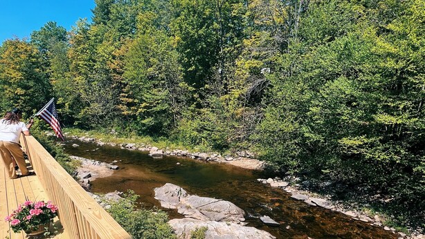 A wooden deck overlooking a river with rocks and dense greenery. - Valley Craft Ales (Wilmington)