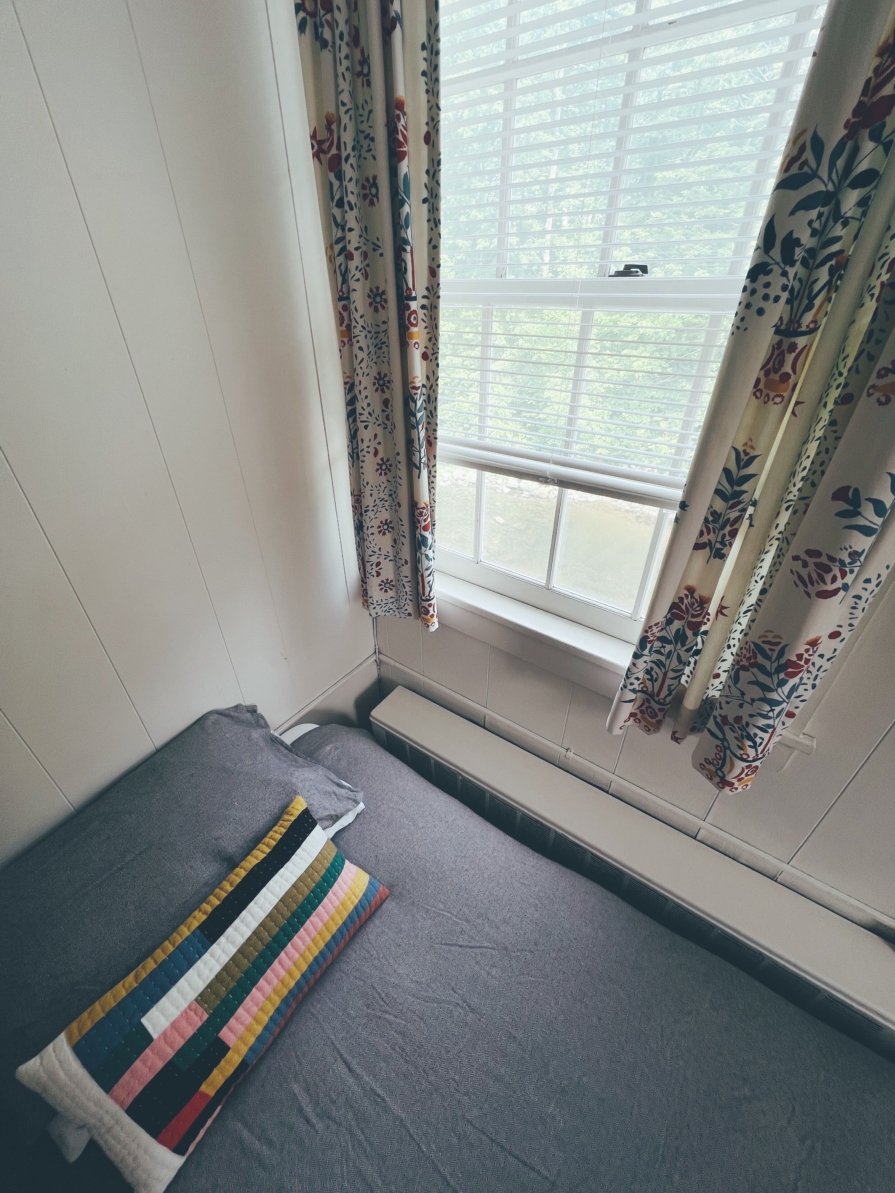 Una habitación pequeña con una cama individual, una ventana con cortinas florales y un radiador.
