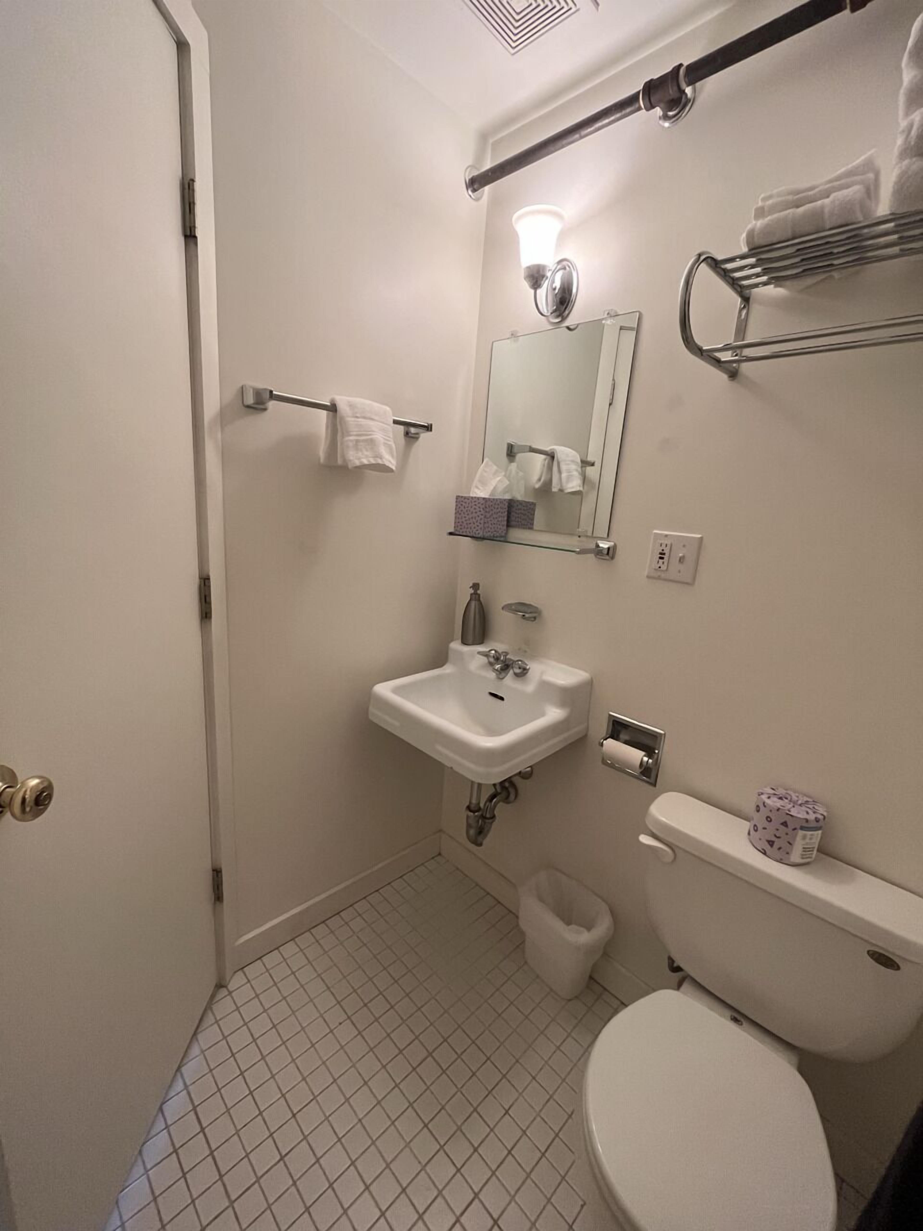 A small, clean bathroom with a toilet, sink, and mirror. There is a towel rack above the sink and a small trash bin next to the toilet.