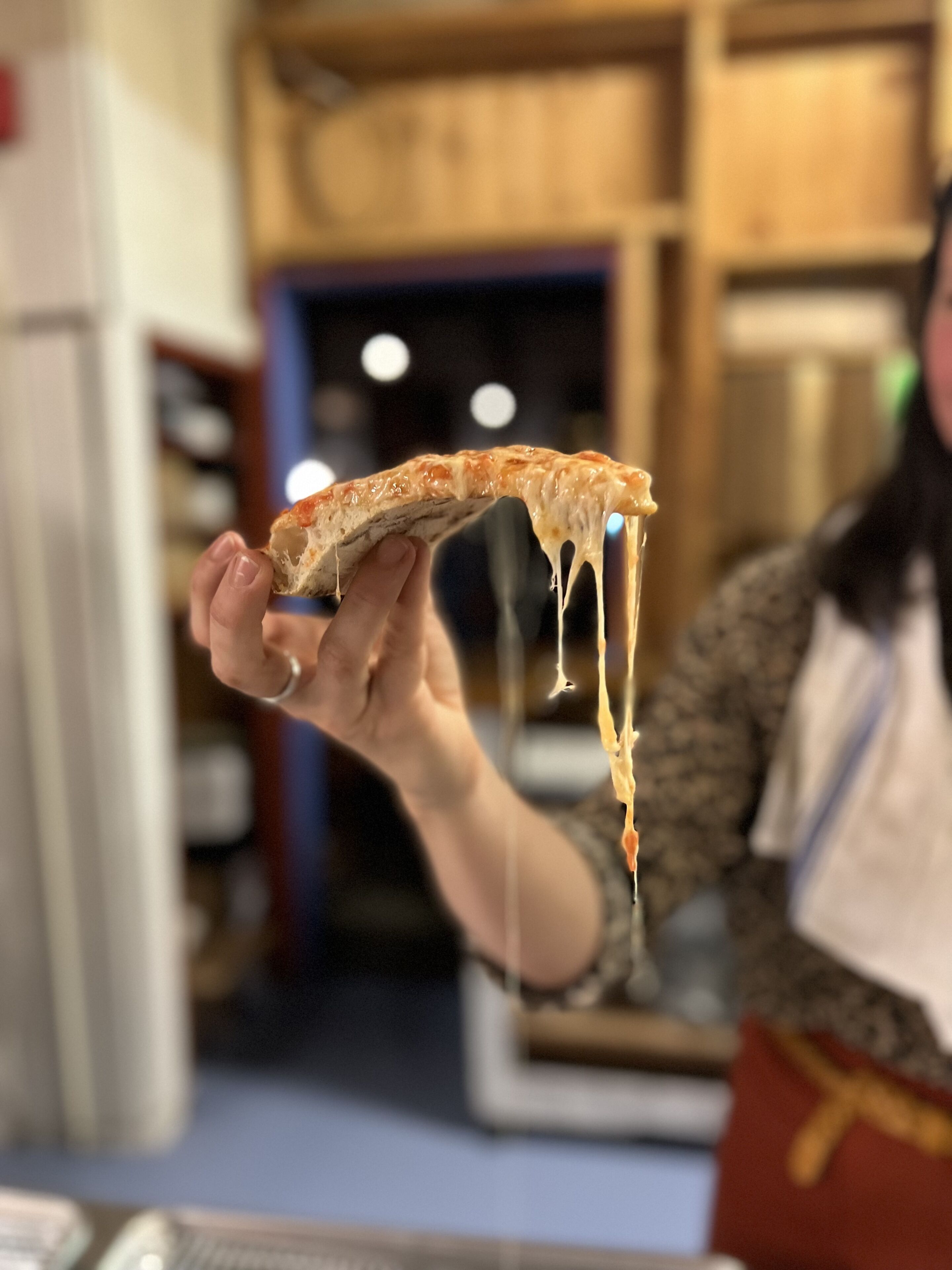 A person holding a slice of pizza with melted cheese, in a kitchen setting.