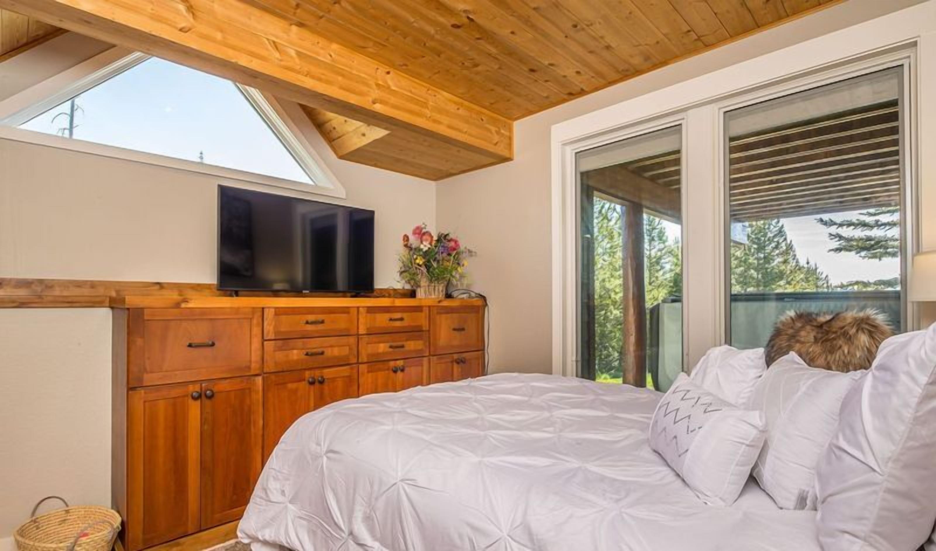 Un dormitorio con techo de madera, una cama grande, un televisor sobre un aparador de madera, y vistas a árboles a través de una puerta corredera.