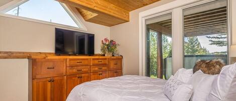 Un dormitorio con techo de madera, una cama grande, un televisor sobre un aparador de madera, y vistas a árboles a través de una puerta corredera.