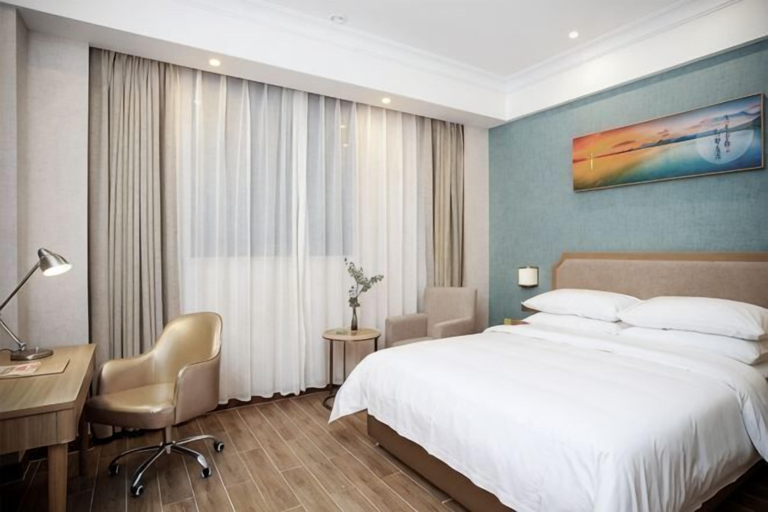 A hotel room with a bed, a desk, a chair, a small table with a lamp, and a painting on the wall.