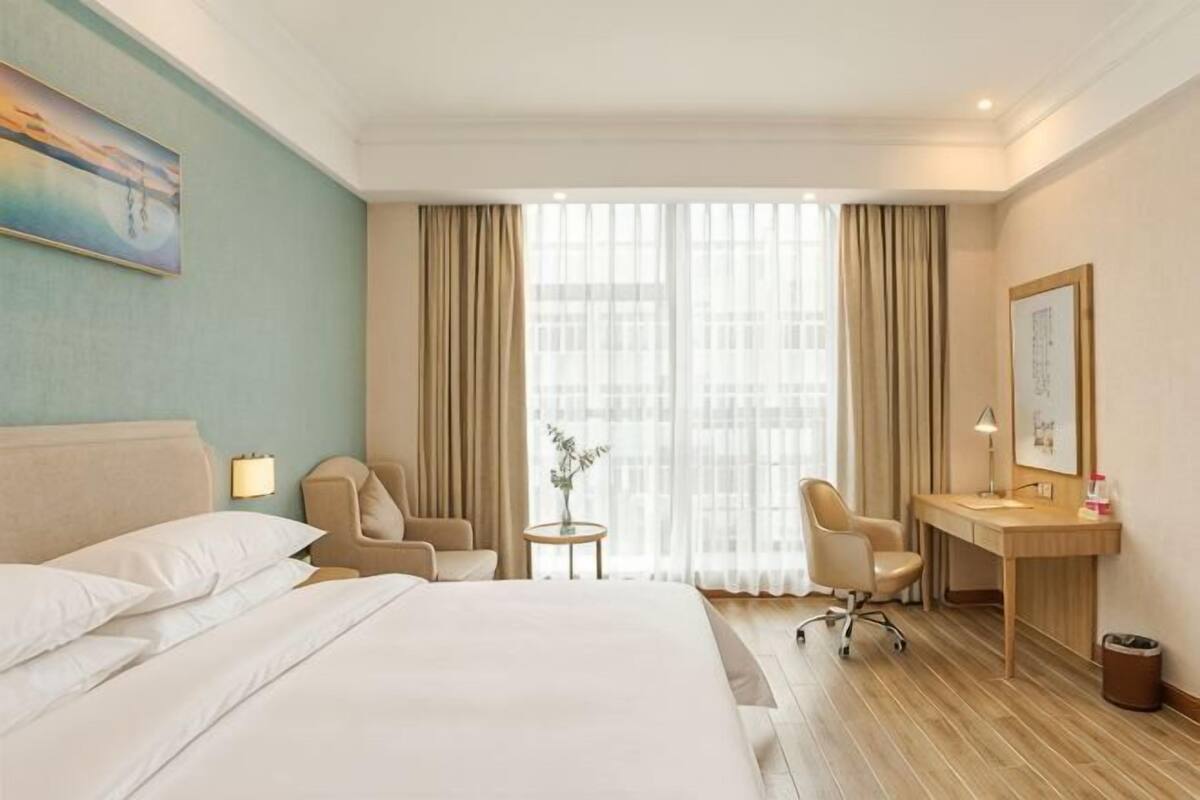 A hotel room with a bed, two chairs, a desk, a small table, and a window with curtains.