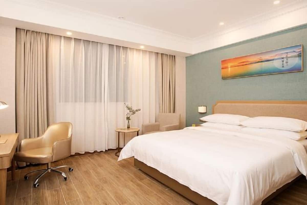 A hotel room with a bed, a desk, a chair, a small table with a vase, and a painting on the wall.