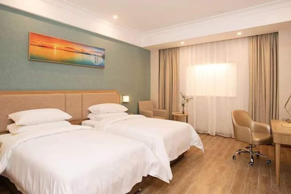 A hotel room with two beds, a desk, a chair, a painting on the wall, and a window with curtains.