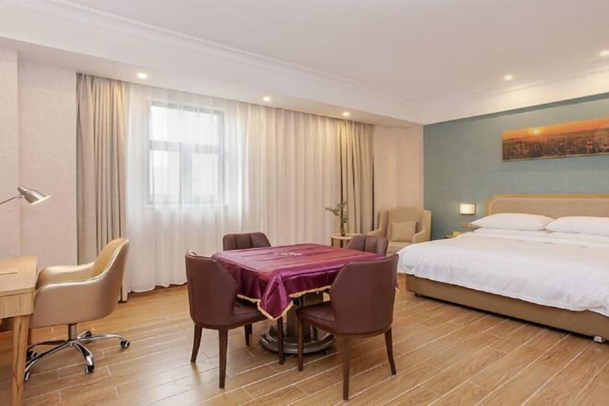 A hotel room with a bed, a desk with a chair, a dining table with chairs, and a window with curtains.