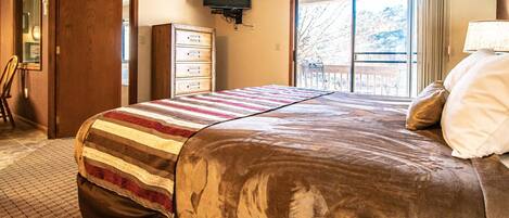 A hotel room with a large bed, a dresser, a television, and a balcony with a view of trees.