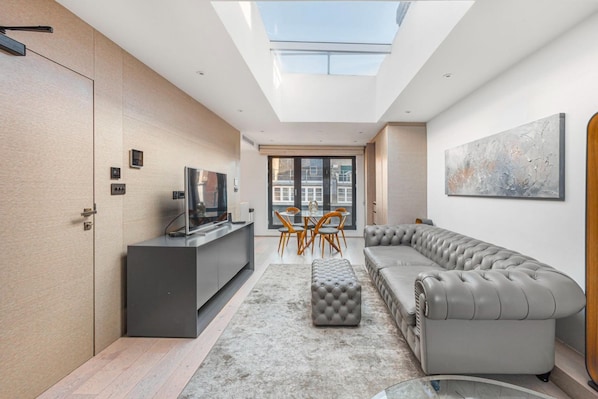 A modern living room with a grey sofa, a glass-top dining table, and a grey ottoman.