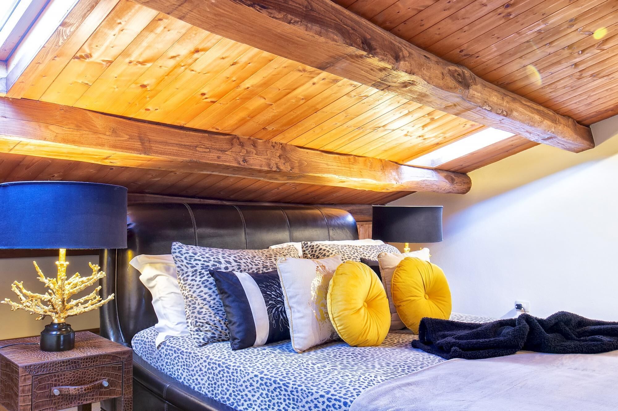 A bedroom with a wooden ceiling, a bed with leopard print bedding, and a nightstand with a lamp and decorative plant.