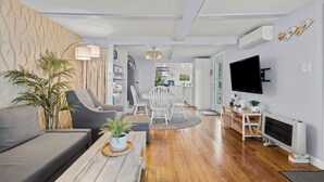 A modern living room with a sofa, coffee table, and TV. There is a dining area with chairs and a table. The room has wooden flooring and a kitchen in the background. - The Dream Location for Your Perfect Vacation  (Branford)