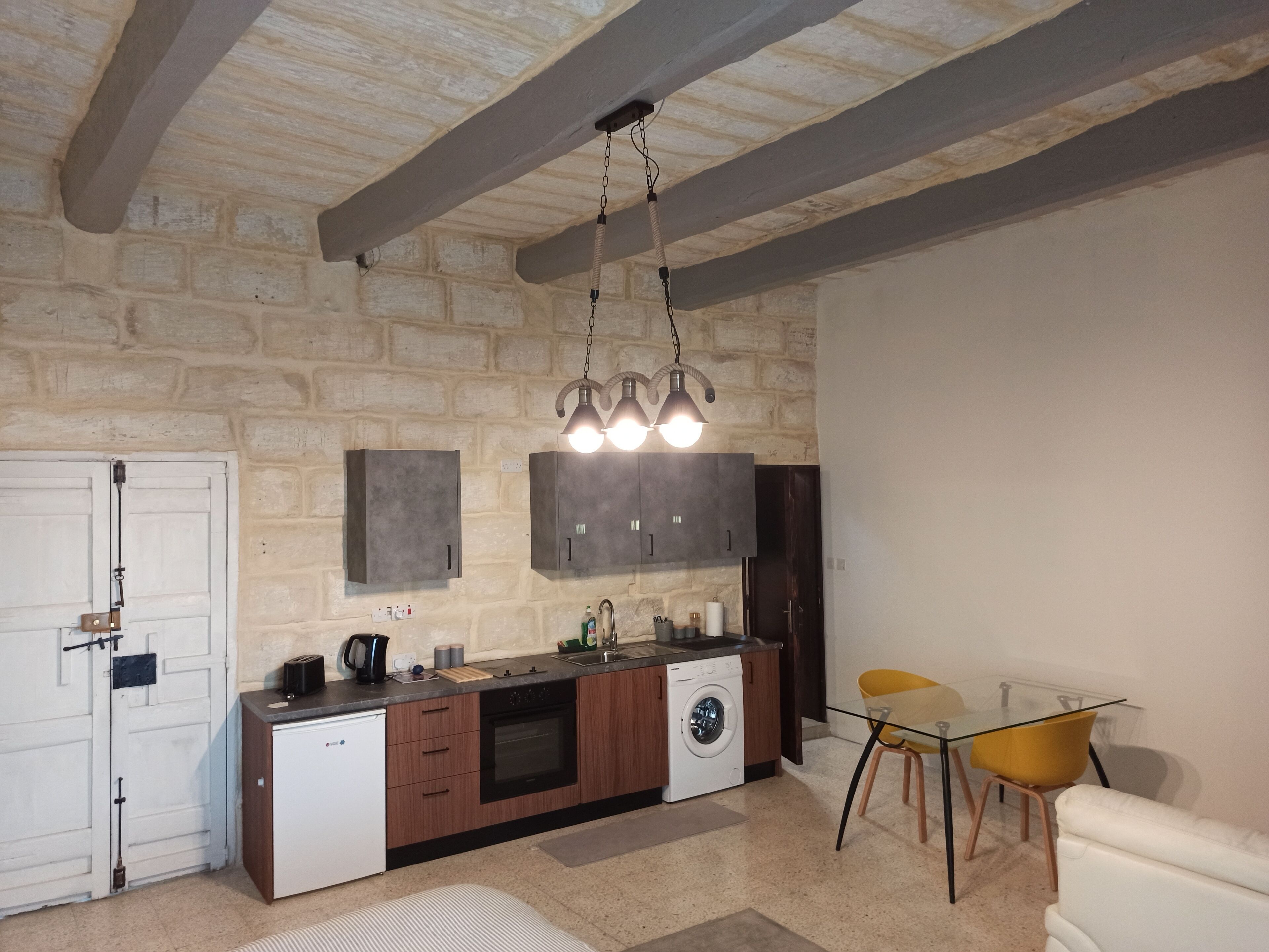 Una cocina compacta con lavadora integrada, un comedor con mesa de vidrio y sillas amarillas, y un fondo de pared de piedra.