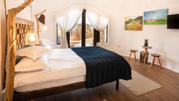 Un dormitorio con cabecera de madera, cama con ropa de cama blanca y oscura, una lámpara de noche, un ventanal con cortinas y dos cuadros en la pared.